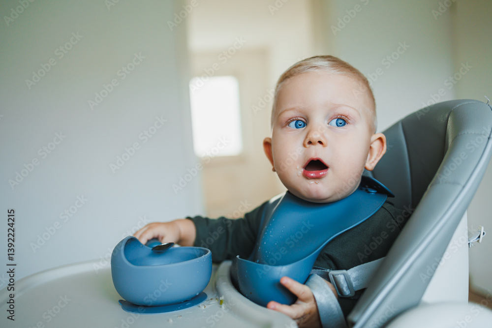 Toddler boy in casual clothes sits at the table in a highchair and eats himself with a spoon. The child learns to eat independently. Healthy nutrition for children. Baby food