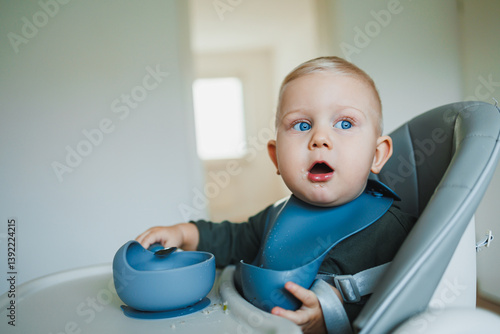 Obraz na płótnie Toddler boy in casual clothes sits at the table in a highchair and eats himself with a spoon. The child learns to eat independently. Healthy nutrition for children. Baby food w salonie