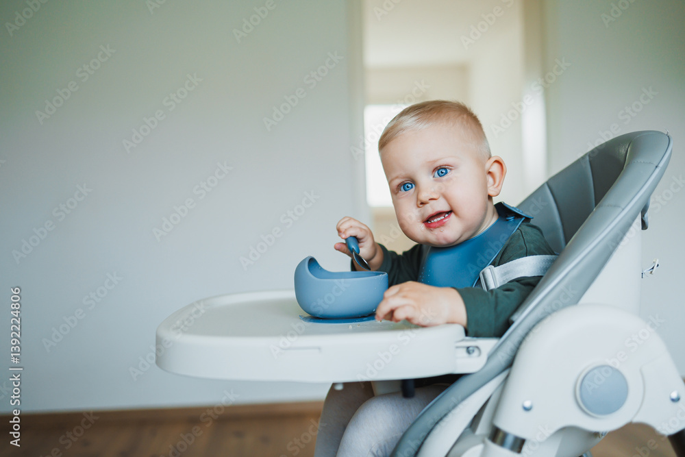 Toddler boy in casual clothes sits at the table in a highchair and eats himself with a spoon. The child learns to eat independently. Healthy nutrition for children. Baby food