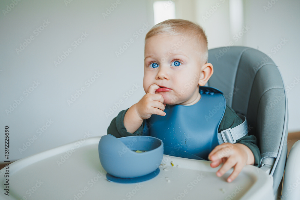 Blue-eyed toddler boy sitting on a chair with a silicone apron eating independently. Toddler boy learning to eat independently. Safe utensils for feeding children.
