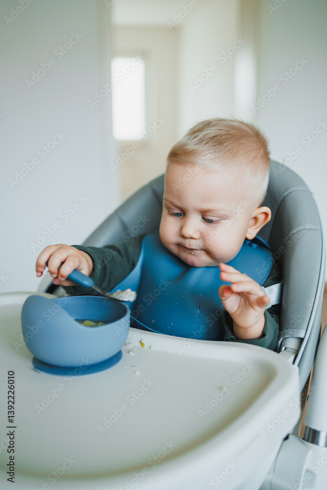 Blue-eyed toddler boy sitting on a chair with a silicone apron eating independently. Toddler boy learning to eat independently. Safe utensils for feeding children.