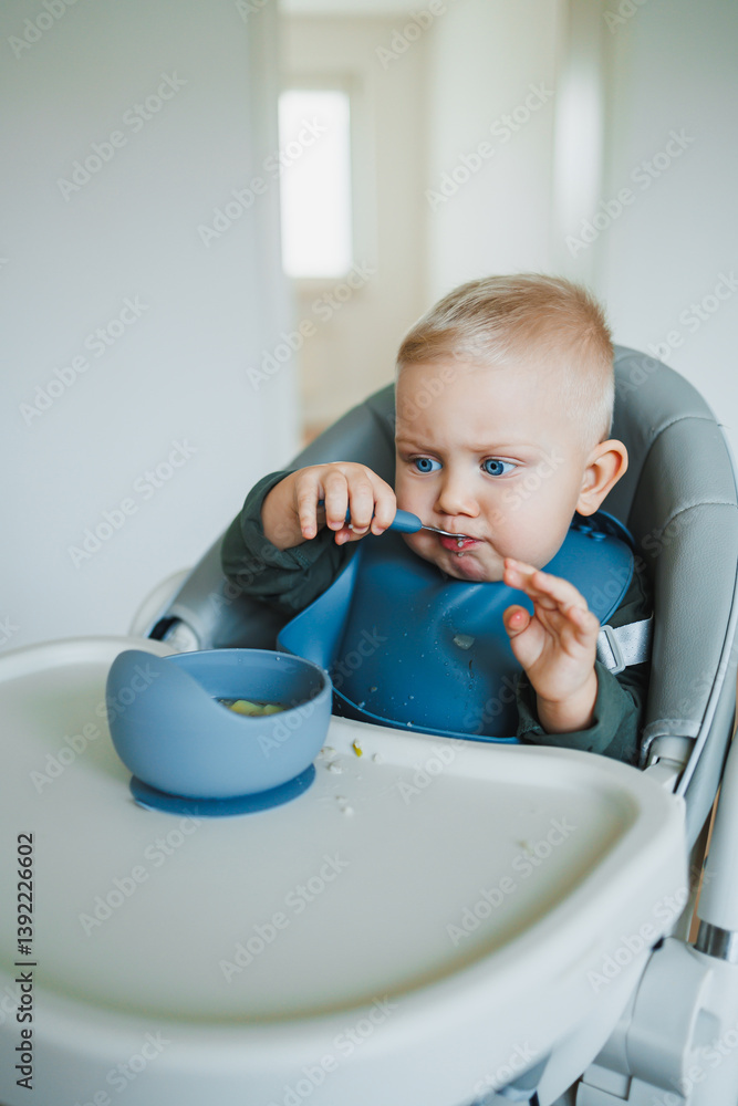 Blue-eyed toddler boy sitting on a chair with a silicone apron eating independently. Toddler boy learning to eat independently. Safe utensils for feeding children.