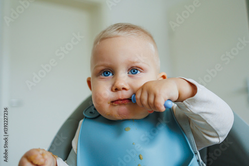 Obraz na płótnie Toddler boy in casual clothes sits at the table in a highchair and eats himself with a spoon. The child learns to eat independently. Healthy nutrition for children. Baby food w salonie