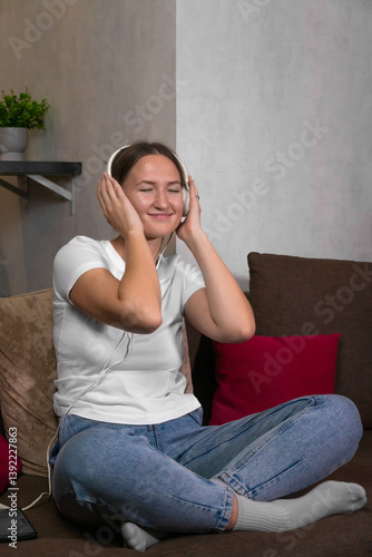 Obraz na płótnie Happy young woman enjoying music through headphones, sitting comfortably on a sofa with her eyes closed, embracing the rhythm and melody. Vertical photo w salonie