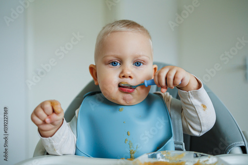 Obraz na płótnie Toddler boy in casual clothes sits at the table in a highchair and eats himself with a spoon. The child learns to eat independently. Healthy nutrition for children. Baby food w salonie