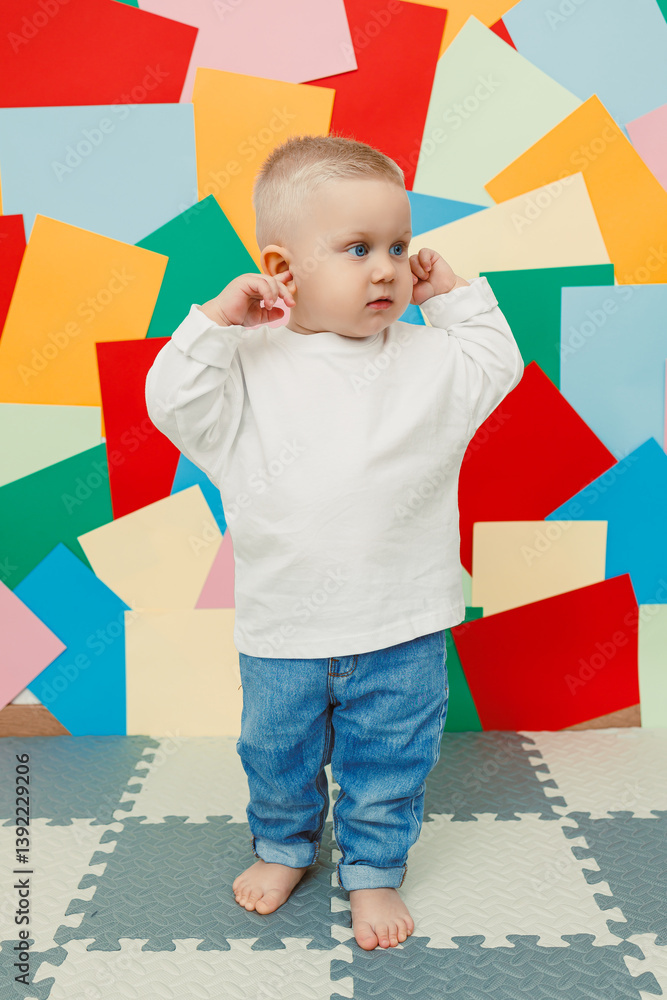Happy little toddler in a white t-shirt and jeans stands on a studio background and covers his ears, expressing joy and curiosity. Stylish toddler. Concept of parenthood, natural beauty, love and care