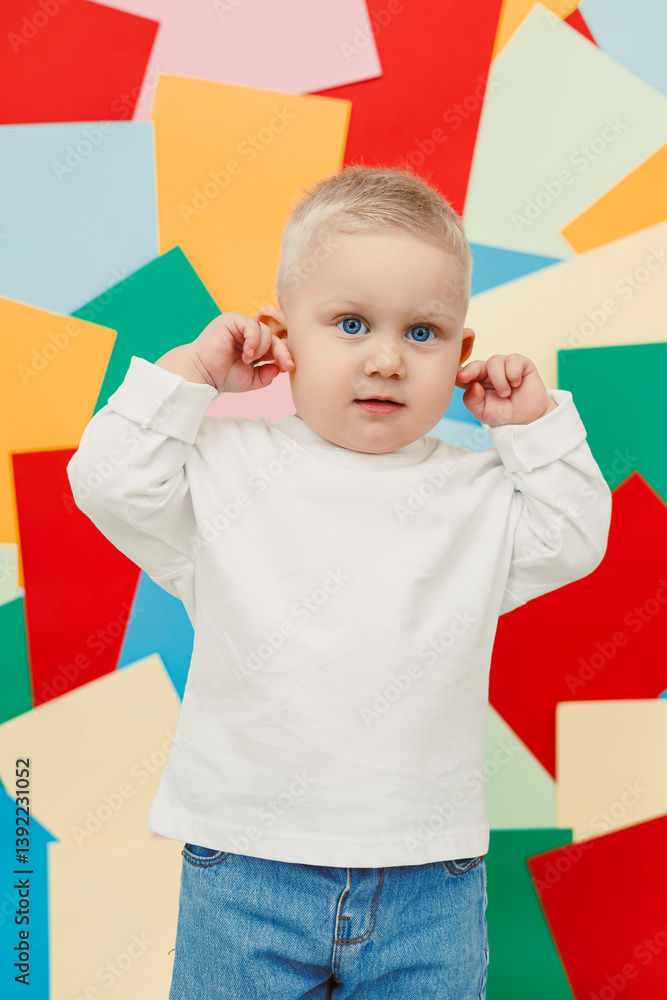 Happy little toddler in a white t-shirt and jeans stands on a studio background and covers his ears, expressing joy and curiosity. Stylish toddler. Concept of parenthood, natural beauty, love and care