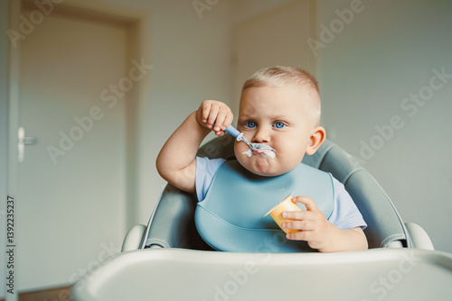 Obraz na płótnie Smiling boy sitting in a high chair, eating yogurt in the kitchen. Healthy food for children. Bio yogurt food for children. Children eating vegetables. Little boy eating dairy products. w salonie