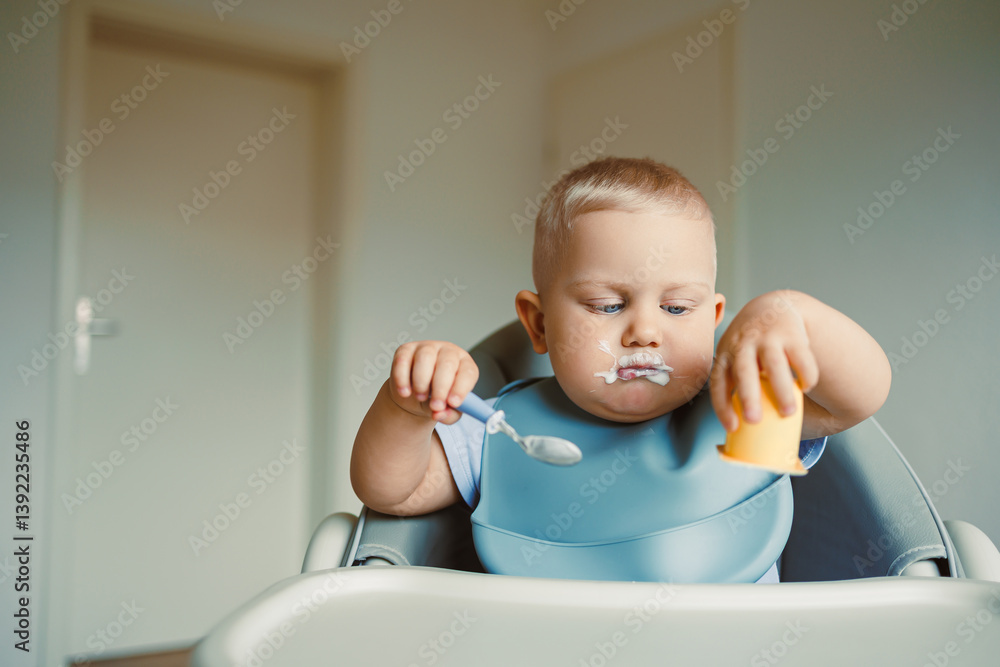 Smiling boy sitting in a high chair, eating yogurt in the kitchen. Healthy food for children. Bio yogurt food for children. Children eating vegetables. Little boy eating dairy products.