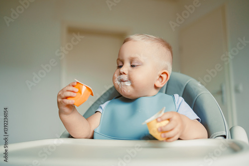 Smiling boy sitting in a high chair, eating yogurt in the kitchen. Healthy food for children. Bio yogurt food for children. Children eating vegetables. Little boy eating dairy products.