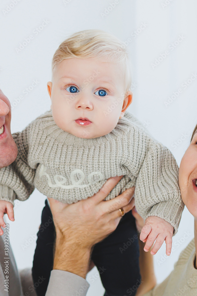 An adorable baby dressed in a cozy sweater and possessing striking blue eyes is lovingly held by their parents. This heartwarming scene takes place in a bright and cheerful indoor setting