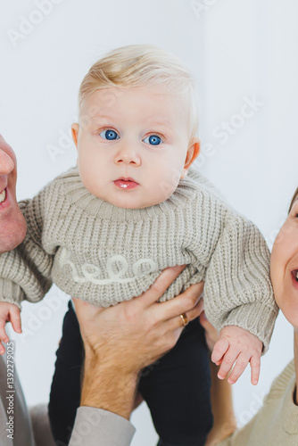 Obraz na płótnie An adorable baby dressed in a cozy sweater and possessing striking blue eyes is lovingly held by their parents. This heartwarming scene takes place in a bright and cheerful indoor setting w salonie