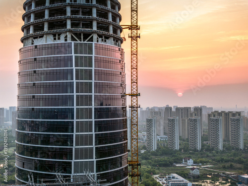 Fotografie Modern circular skyscraper building under construction with crane against cityscape residential area background at sunset