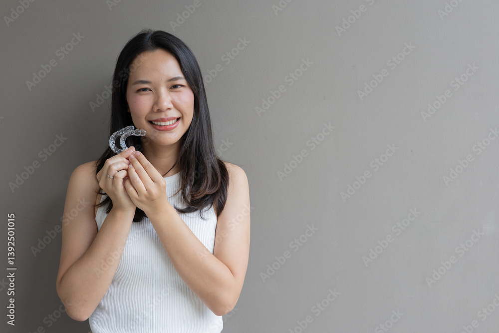 close up young asian beautiful woman smiling with hand holding dental aligner retainer (invisible) on grey background at dental clinic for beautiful teeth treatment course concept close up young asian beautiful woman smiling with hand holding dental aligner retainer (invisible) on grey background at dental clinic for beautiful teeth treatment course concept