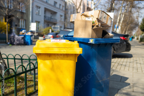 Obraz na płótnie Two trash bins, one yellow and one blue, placed on the sidewalk. The bins are filled with cardboard boxes and waste, surrounded by greenery and urban buildings in the background. A clean street and w salonie