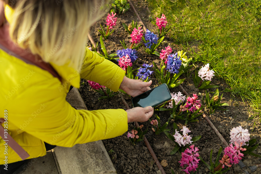Woman in a yellow jacket takes a photo of vibrant flowers in a spring garden using her smartphone. The image captures the beauty of nature and the joy of capturing memories.