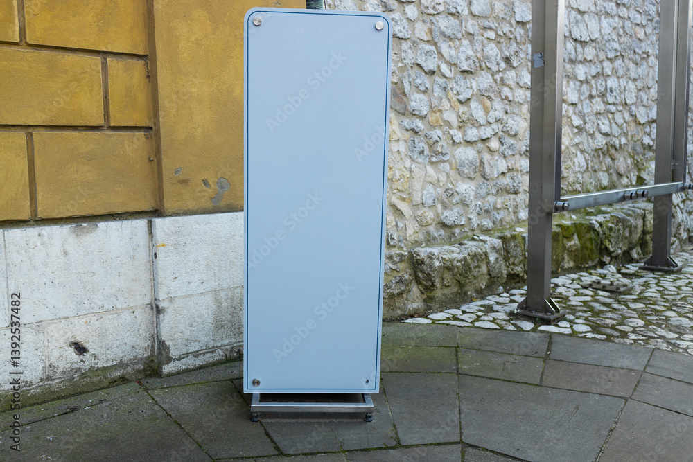 Blank advertising board stand placed at an urban street corner. The simple and clean design creates space for advertisements or promotional content, standing on a cobbled surface against a stone wall.