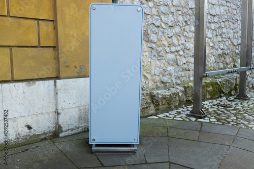 Obraz na płótnie Blank advertising board stand placed at an urban street corner. The simple and clean design creates space for advertisements or promotional content, standing on a cobbled surface against a stone wall. w salonie