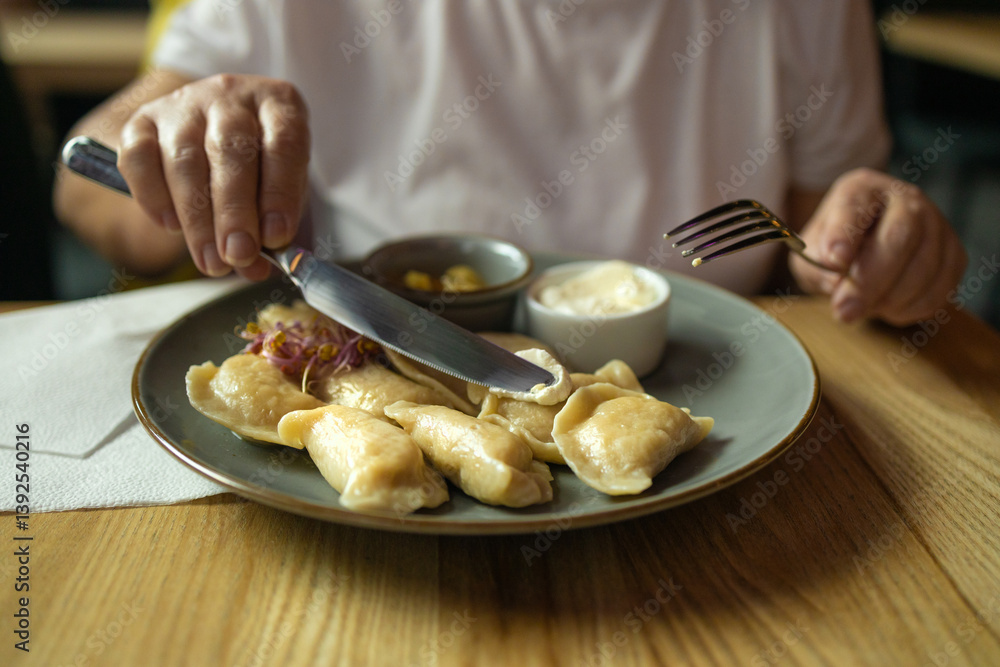 Person is holding a knife and fork, ready to eat a plate of traditional dumplings served with a side of sour cream and another small dish. The scene is in a cozy restaurant, showcasing delicious