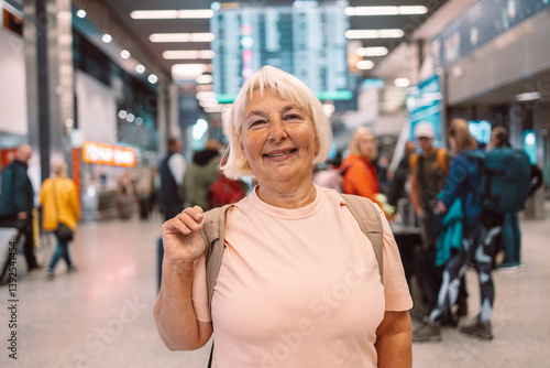 Obraz na płótnie Happy elderly woman in a pink t-shirt with a backpack, smiling at the airport terminal. She ready for her next travel adventure. Ideal for travel, senior lifestyle, and vacation content. w salonie