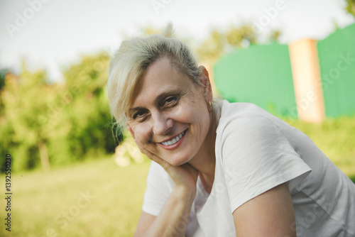 Obraz na płótnie A joyful woman with short blonde hair, wearing a white t-shirt, smiles softly while resting her head on her hand. She sits in a sunny outdoor garden with green trees and a fence in the background. w salonie