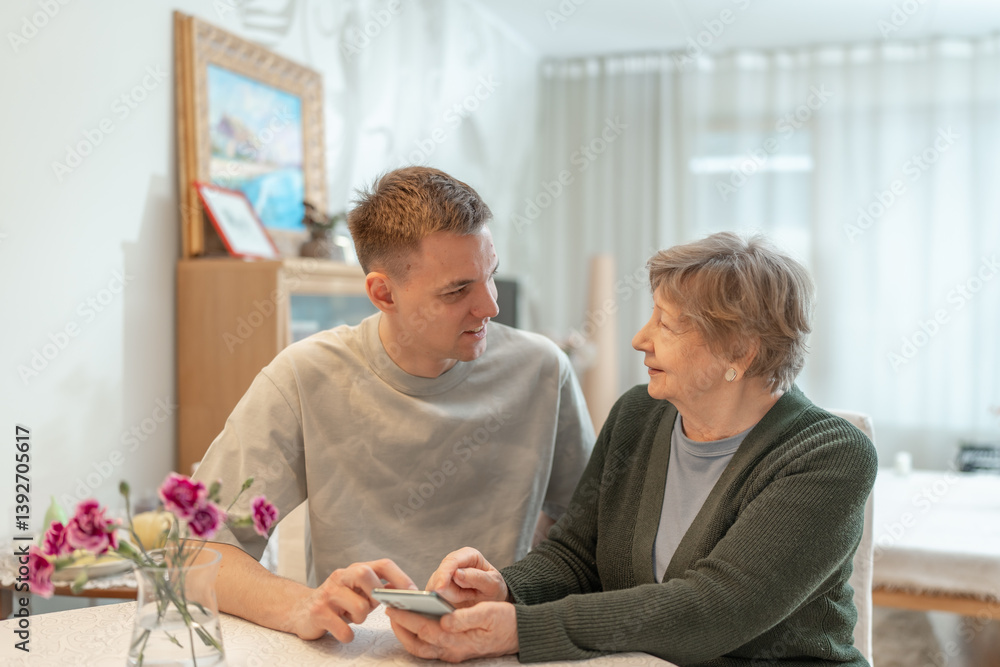 Fototapeta premium Young man helping senior woman with a smartphone at home. Concept of technology, education, and senior digital adaptation. Grandson teaches grandmother how to use digital device. Assisted living.