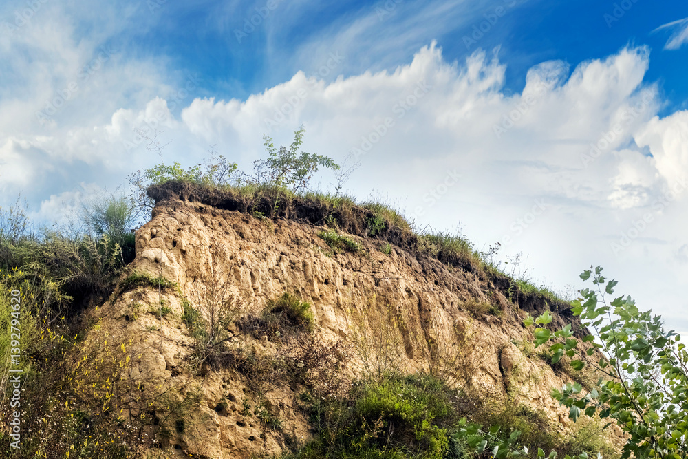 Low angle view of a clay cliff covered with vegetation against a blue sky with clouds