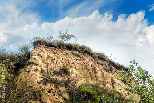 Obraz na płótnie Low angle view of a clay cliff covered with vegetation against a blue sky with clouds w salonie