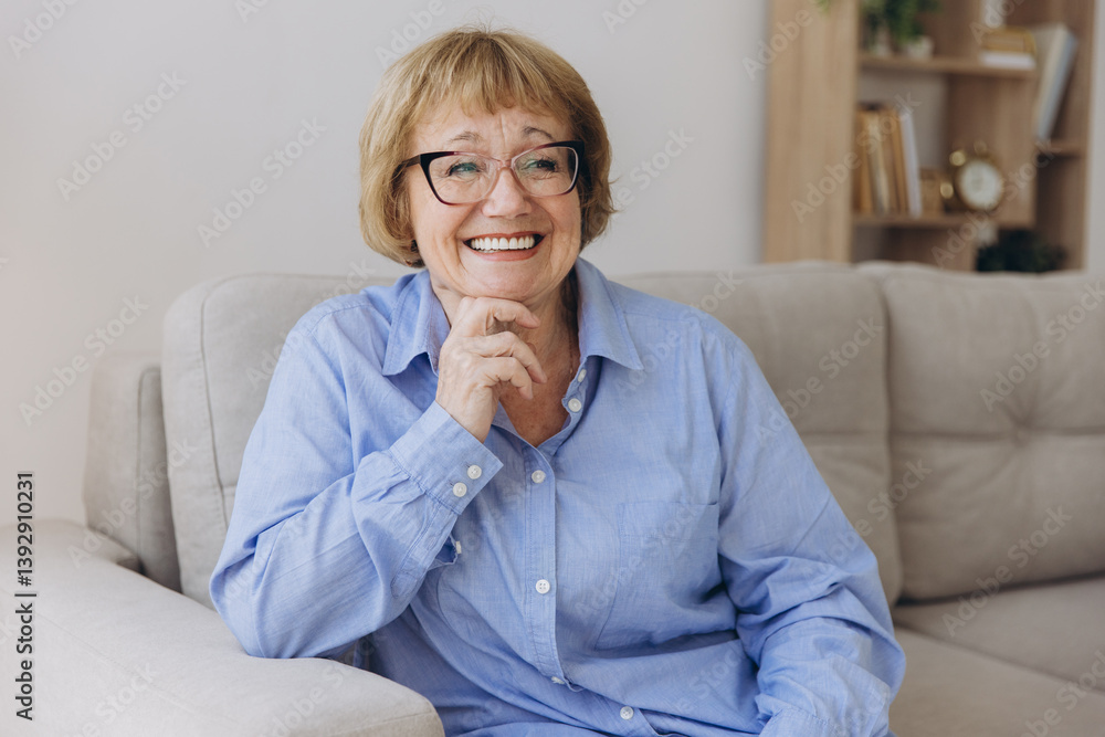 Smiling senior blonde haired woman looking at camera, happy old lady in glasses posing at home indoor, positive single senior retired female sitting on sofa in living room headshot portrait Smiling senior blonde haired woman looking at camera, happy old lady in glasses posing at home indoor, positive single senior retired female sitting on sofa in living room headshot portrait