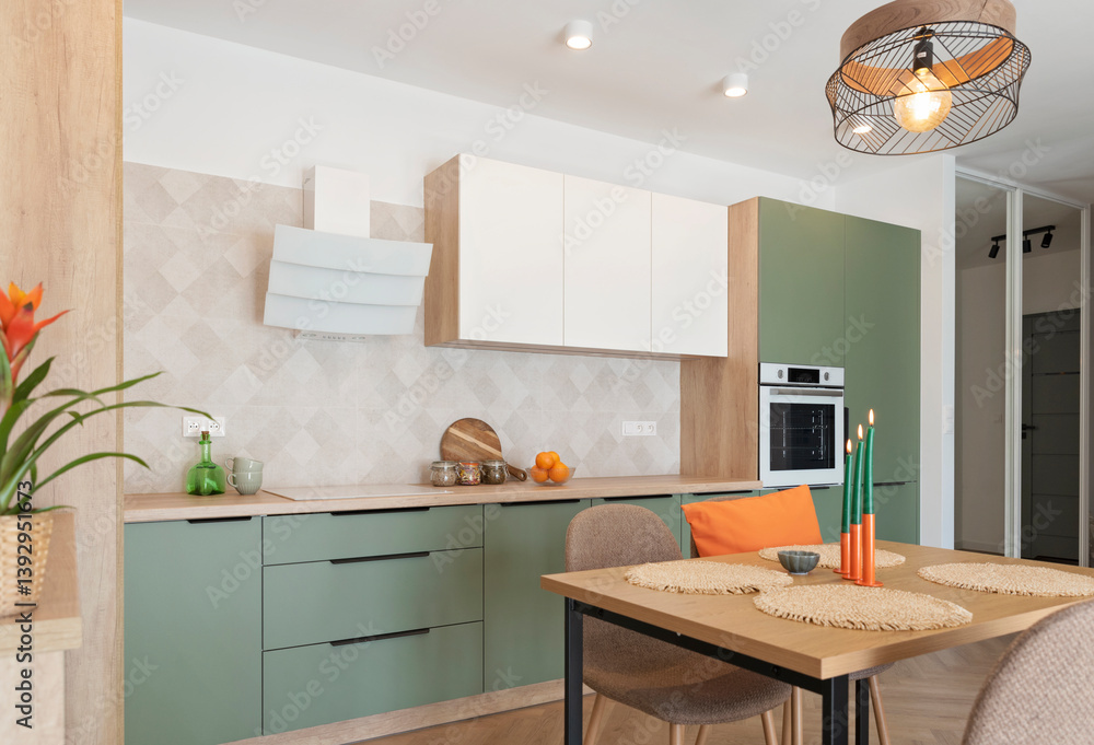 Fototapeta premium Modern kitchen interior with table and chairs. Green and white furniture with cabinet, oven and eaves and orange decoration. Design in new apartment. Real estate.