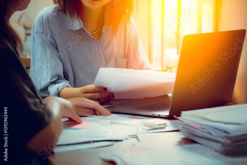 Two individuals collaborate over documents and a laptop, bathed in warm sunlight, suggesting a productive work environment