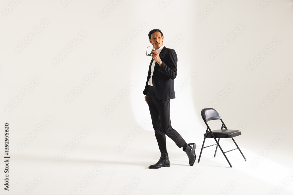 Confident man in suit holding glasses, posing stylishly beside chair in studio Confident man in suit holding glasses, posing stylishly beside chair in studio