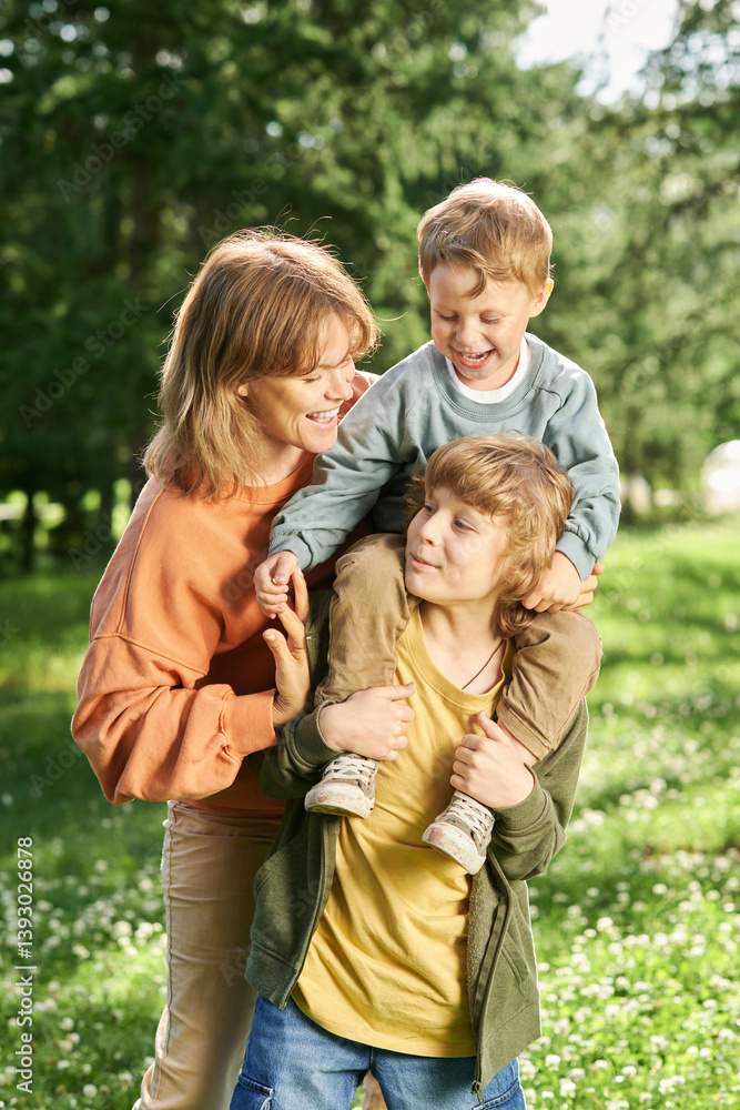 Fototapeta premium Vertical portrait of family enjoying time together in green park young boy holding little brother on shoulders with mother helping