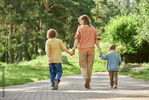 Full length back view of mother with two boys holding hands and walking away from camera in green park copy space