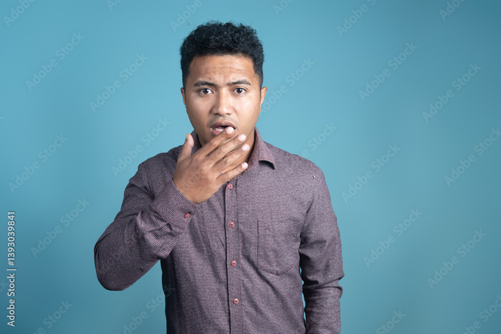 Young man in a button-up shirt looking surprised with hand covering his mouth, standing against a plain blue background. Emotion concept, reaction expression, studio shot. Young man in a button-up shirt looking surprised with hand covering his mouth, standing against a plain blue background. Emotion concept, reaction expression, studio shot.