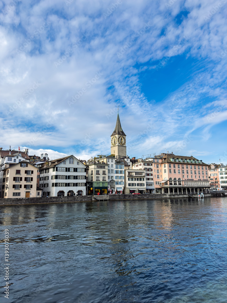Fototapeta premium Historic Zurich Waterfront with St. Peter Church Tower and Limmat River.Zurich Cityscape: Medieval Architecture along Limmat River under Blue Sky.