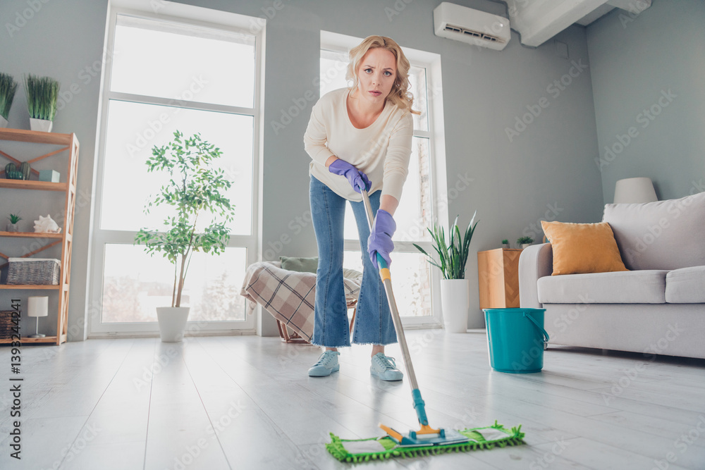 Young blonde woman mopping living room floor in a stylish comfortable home interior with sunlight streaming through large windows Young blonde woman mopping living room floor in a stylish comfortable home interior with sunlight streaming through large windows