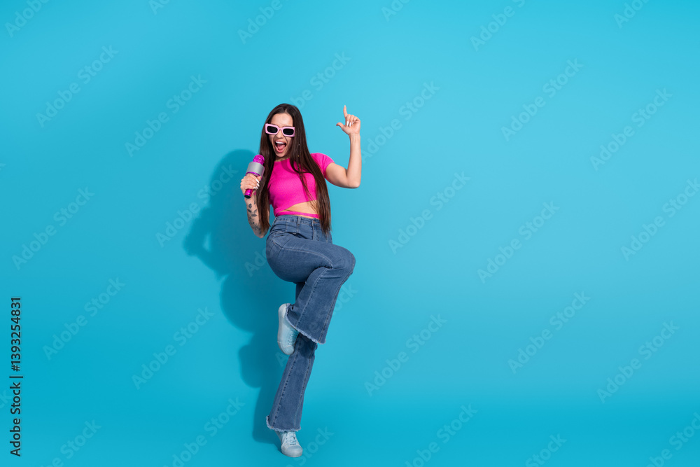 Obraz premium Young woman in stylish pink top and jeans posing joyfully with sunglasses on a bright blue background, expressing fun and trendy lifestyle.
