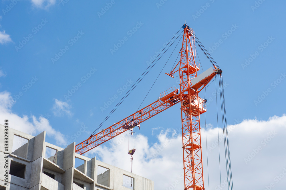 Tall orange construction crane towers over a building site, against a backdrop of a bright blue sky with clouds. Tall orange construction crane towers over a building site, against a backdrop of a bright blue sky with clouds.