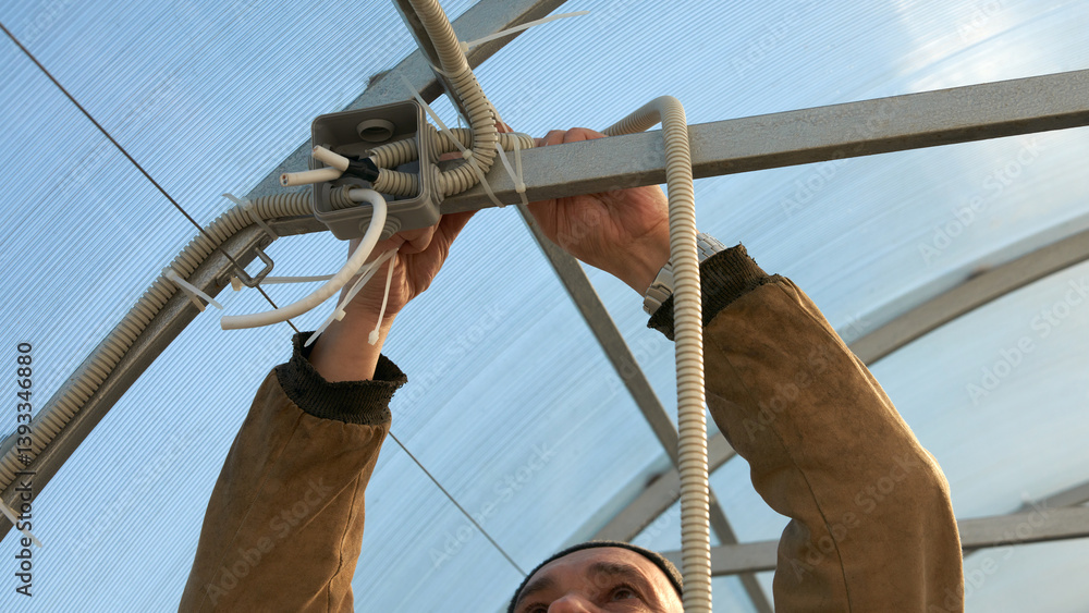 Electrical wiring in a polycarbonate greenhouse. A man brings the electric cable laid in corrugated tube to the infrared heater.
