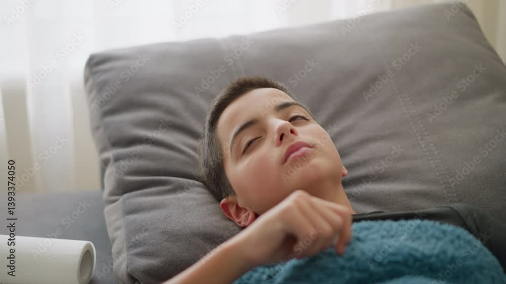 Close-up of sick boy resting on couch covered with blue blanket, coughing, adjusting position while feeling unwell with light streaming through window Close-up of sick boy resting on couch covered with blue blanket, coughing, adjusting position while feeling unwell with light streaming through window