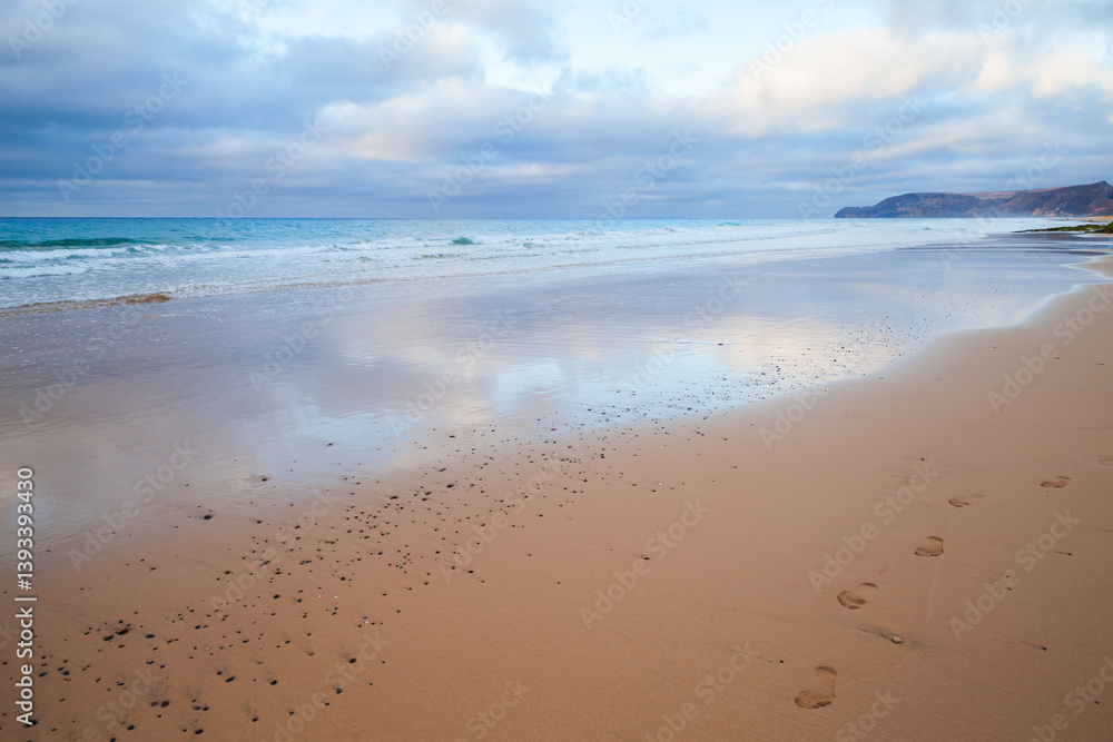 Serene beach scene with gentle ocean waves, cloudy skies, and footprints