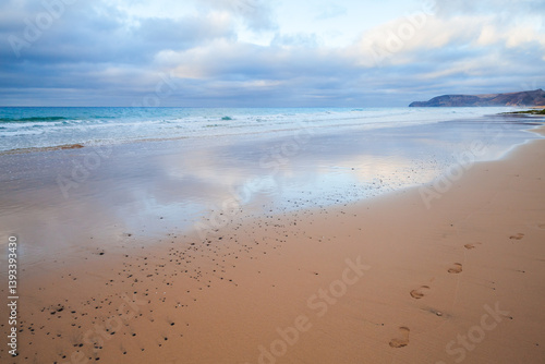 Obraz na płótnie Serene beach scene with gentle ocean waves, cloudy skies, and footprints w salonie