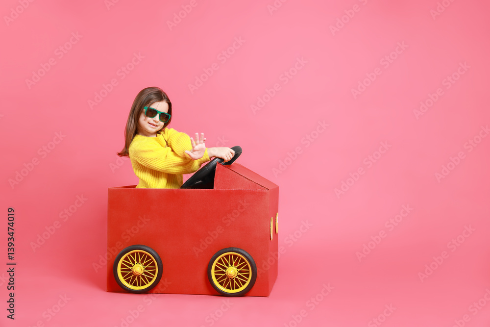 Little girl in sunglasses driving car made with cardboard on red background. Space for text