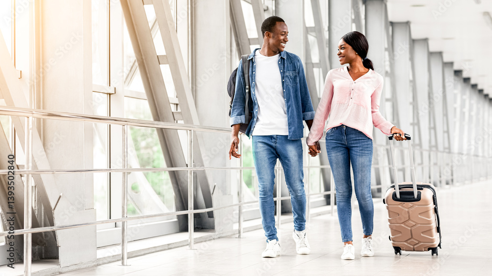 Fototapeta premium Journey Together. Smiling Black Spouses Walking With Luggage In Airport Terminal, Romantic African American Couple Holding Hands While Going To Gate, Having Honeymoon Or Vacation Trip, Free Space