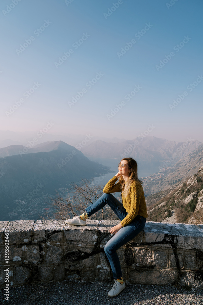 Naklejka premium Smiling girl sits on the curb of the observation deck over the Bay of Kotor. Montenegro