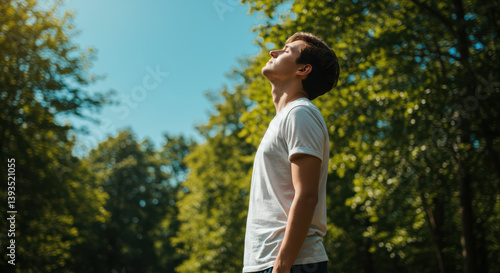 Young man breathes fresh air in natural atmosphere. Adult guy tilts head up and closes eyes in forest and blue sky. He enjoys vacation, meditates, feels calm.