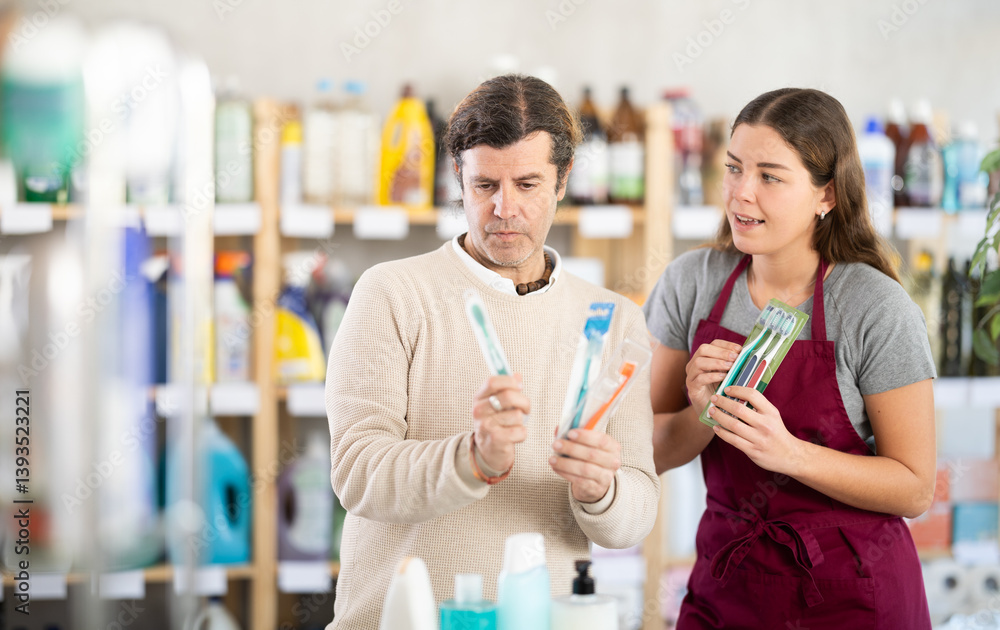 Interested middle-aged customer carefully examining packaged toothbrushes while polite young female sales assistant offering additional options in store selling hygiene products