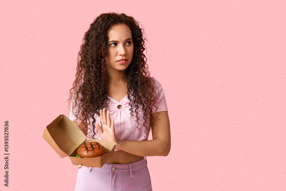 Fototapeta premium Young African-American woman showing stop gesture against burger on pink background. Diet concept
