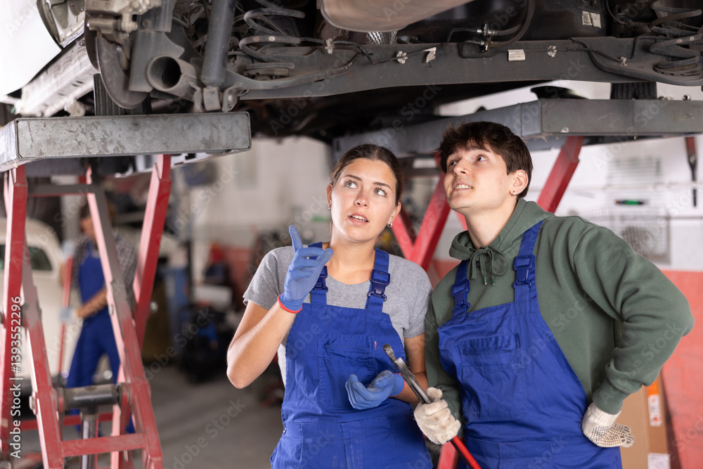 Two focused young auto mechanics, girl and guy, in blue overalls working together, inspecting car lifted on hydraulic lift in vehicle repair shop and discussing reason for breakdown